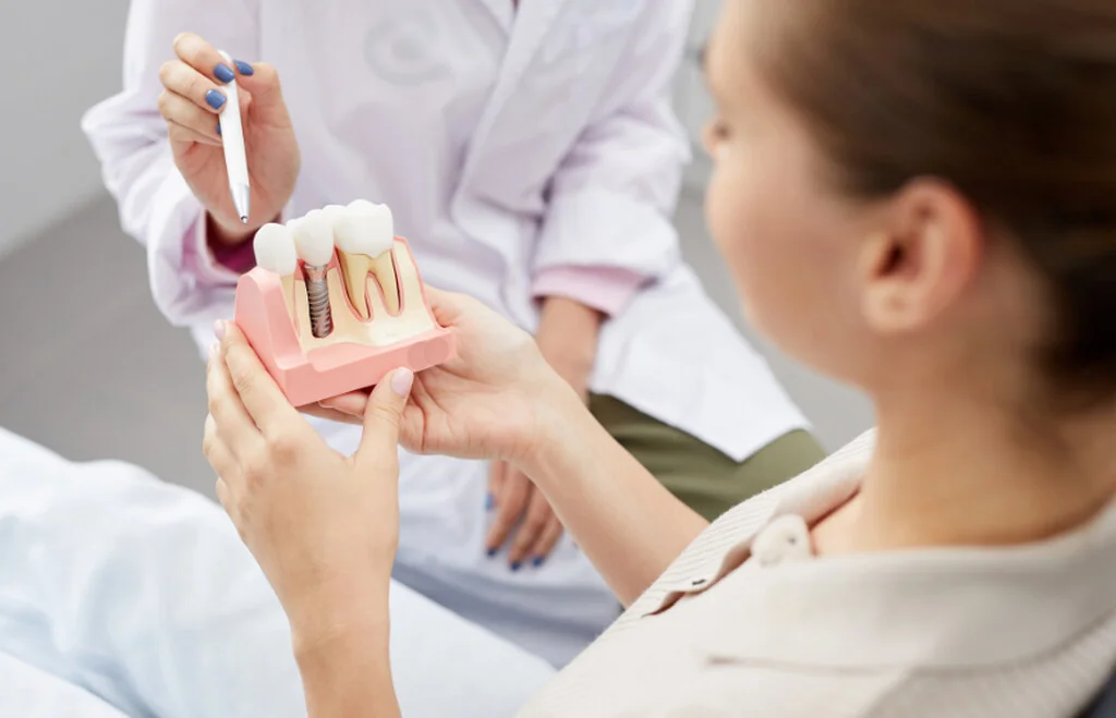 Pani stomatolog tłumacząca pacjentce na czym polega leczenie implantologiczne. Pacjentka trzyma w ręce model z implantem zęba.