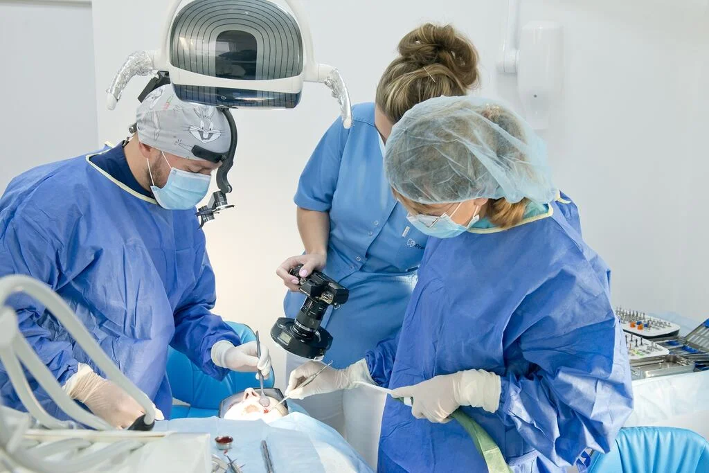 Zabieg Implantologiczny w PrecisDENT - Centrum Implantologii i Stomatologii Mikroskopowej. Lekarz wprowadza implant zęba, jedna asystentka trzyma ssak i pomaga odsłaniać pole zabiegowe. Druga asystentka wykonuje zdjęcie do dokumentacji medycznej zabiegu.