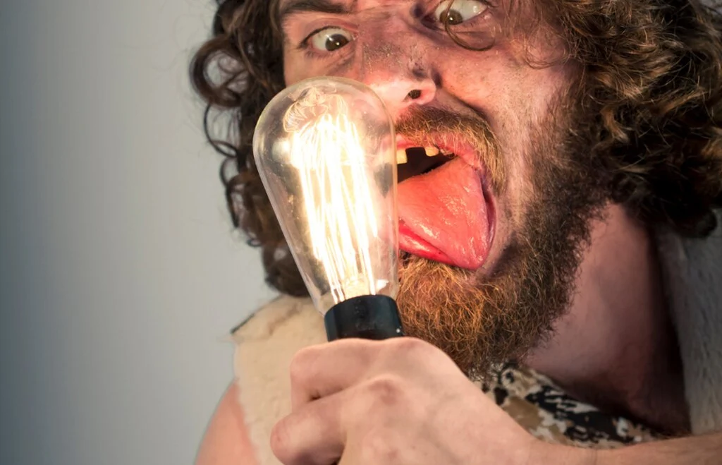Człowiek pierwotny z zezem, bez zęba przedniego, z wystawiony w bok językiem patrzy na świecącą żarówkę i sprawia wrażenie głupiego