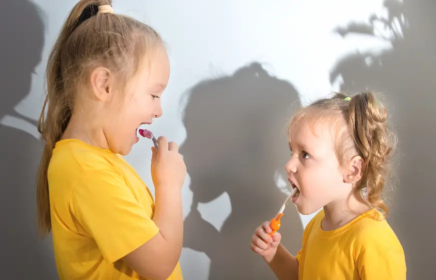 Dziewczynki w żółtych bluzkach myją zęby szczoteczką do zębów patrząc na siebie