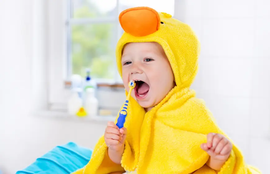 dziecko w żółtym ręczniku z kapturem w kształcie kaczki na głowie z radością myje zęby szczoteczką dla dzieci w żółto-niebieskim kolorze