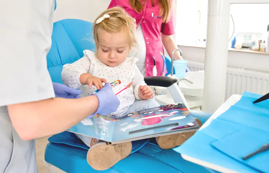 Pierwsza wizyta dziecka w PrecisDENT - Centrum Implantologii i Stomatologii Mikroskopowej w Jaśle. Dziewczynka koloruje wodą specjalną książeczkę dla dzieci na fotelu dentystycznym w obecności Pani stomatolog i asystentki stomatologicznej