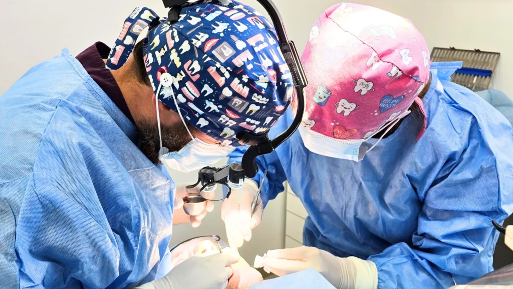 Zabieg stomatologiczny – implantolog i asystentka stomatologiczna w ochronnych czepkach i lupach zabiegowych wykonuje precyzyjną operację implantologiczną w gabinecie.