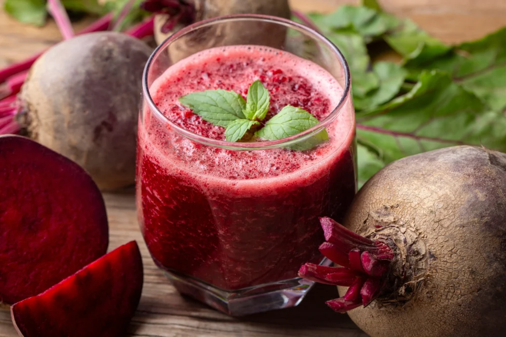 Szklanka czerwonego soku z buraka z listkami mięty i słomką, stojąca obok całych i pokrojonych buraków na drewnianym stole.