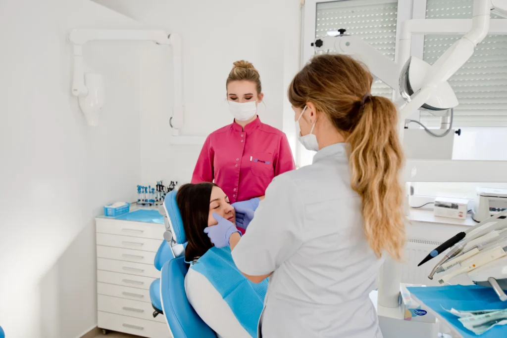 Fotografia przedstawia wnętrze nowoczesnego, jasnego gabinetu dentystycznego. Na niebieskim fotelu stomatologicznym siedzi pacjentka z zamkniętymi oczami, ubrana w białą bluzkę i przykryta niebieskim śliniakiem ochronnym. Stoi nad nią dentystka Joanna Skocz w szarym kitlu i maseczce ochronnej, która bada palpacyjnie policzek pacjentki dłońmi w fioletowych rękawiczkach. W tle, za fotelem, widoczna jest asystentka stomatologiczna w różowym kitlu i maseczce, patrząca na przebieg badania. Tło gabinetu jest czyste, ze sterylną białą szafką medyczną z szufladami, na której stoją materiały stomatologiczne, oraz profesjonalnym ramieniem lampy dentystycznej. Światło jest jasne i naturalne.